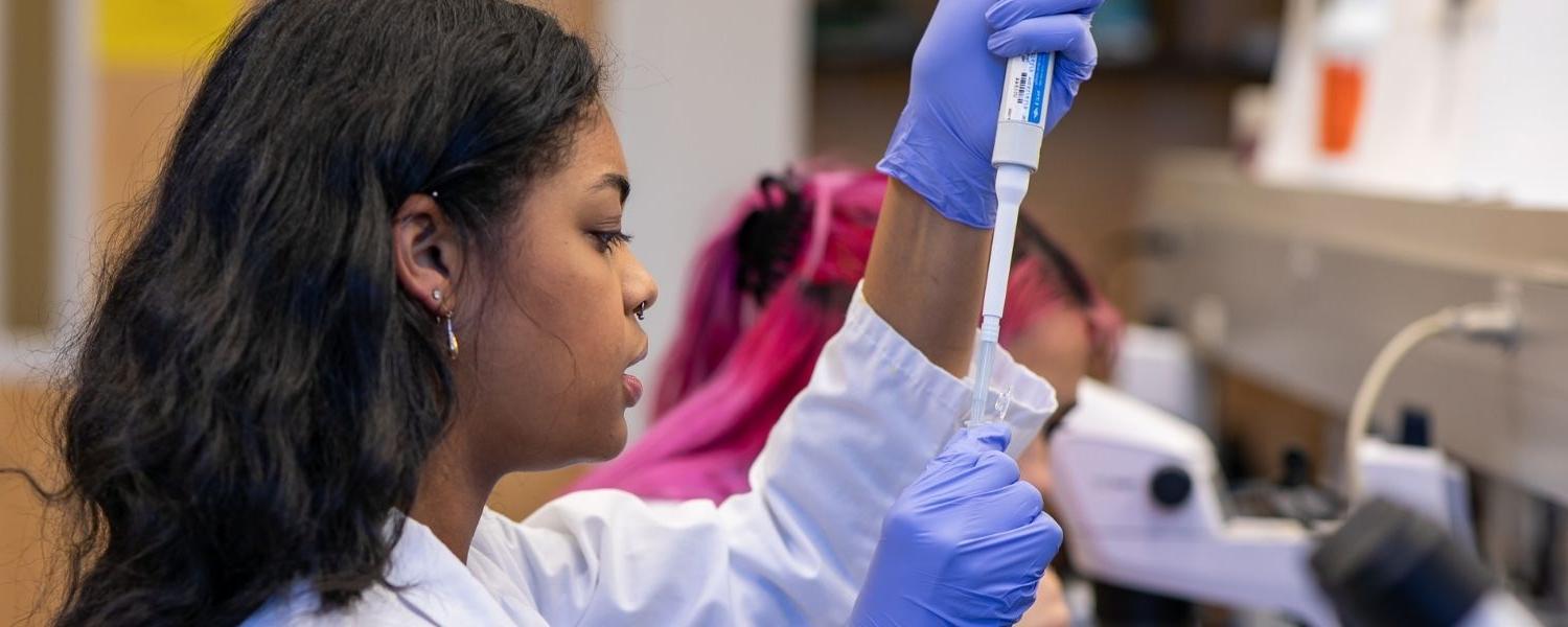 An SPU student works in a lab, adding a liquid to a test tube, as her fellow student works in the background.